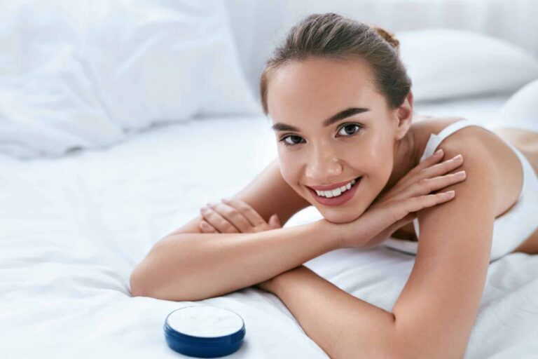 A woman smiles while lying on a white bed, aware of the benefits antioxidants provide for her skin. She takes diligent care to enjoy these effects.