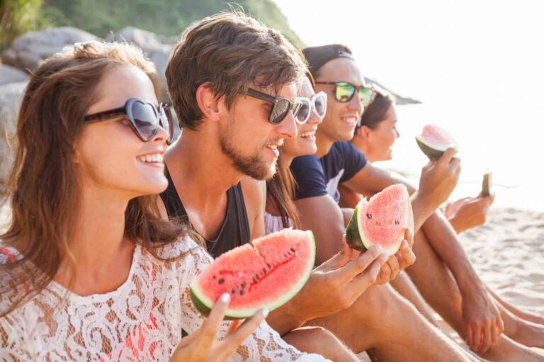 Happy young friends eating watermelon on the beach, unknowingly benefiting from its ability to promote younger-looking skin.
