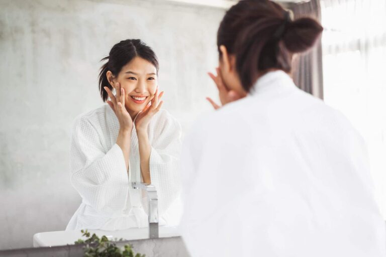 A smiling woman in a white robe touches her face in front of a mirror, demonstrating a Korean skincare routine.