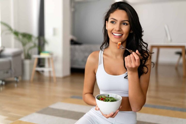 A beautiful woman with dark hair, dressed in white workout clothes, enjoys a bowl of healthy salad, knowing how to get glowing skin naturally.