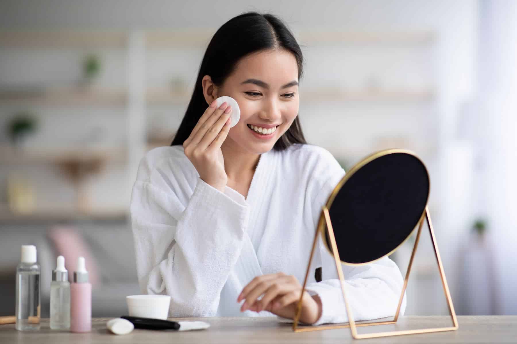 A woman in a white robe smiles while using a cotton pad to remove makeup at a mirror, demonstrating a step in the 10 step Korean skincare routine.