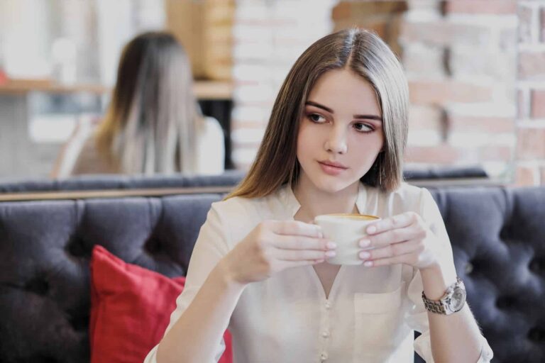 An attractive woman in a white blouse sits alone at a café with a cappuccino, her confidence standing out as one of her most appealing traits.
