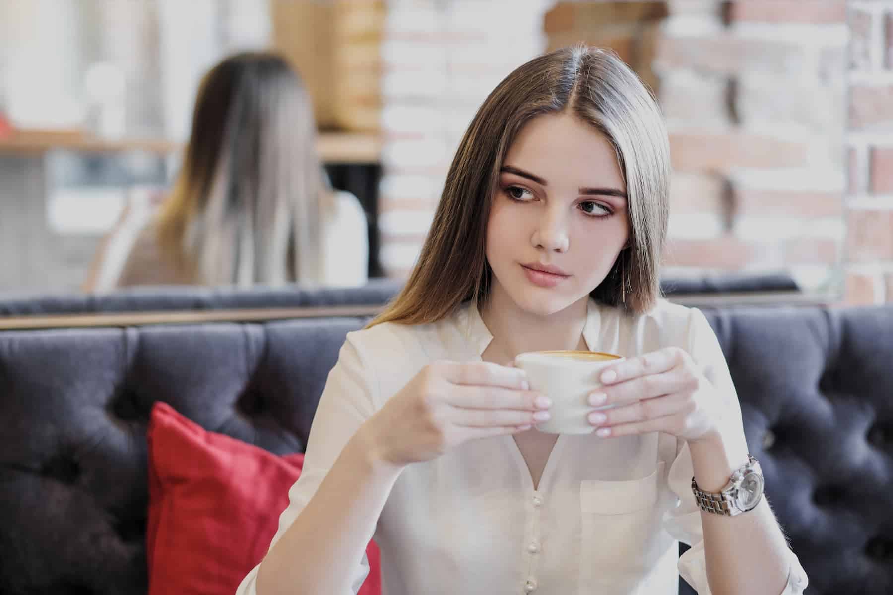An attractive woman in a white blouse sits alone at a café with a cappuccino.