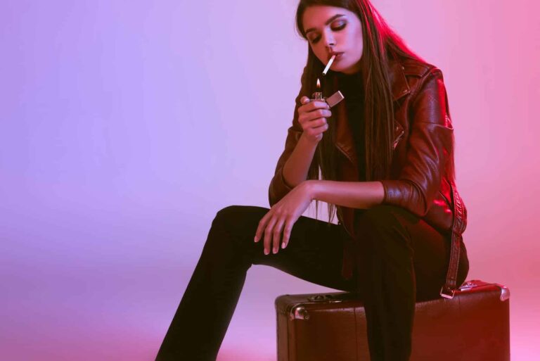 A woman in a leather jacket sits on a suitcase, lighting a cigarette against a violet background, unaware of how smoking affects her skin.