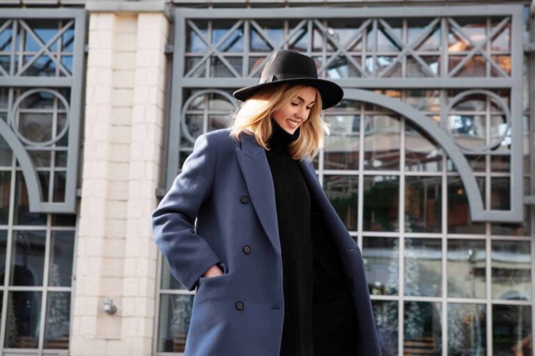 A stylish woman outdoors in a black hat, turtleneck dress, and blue overcoat, showcasing elegance and best beauty tips for a chic look.