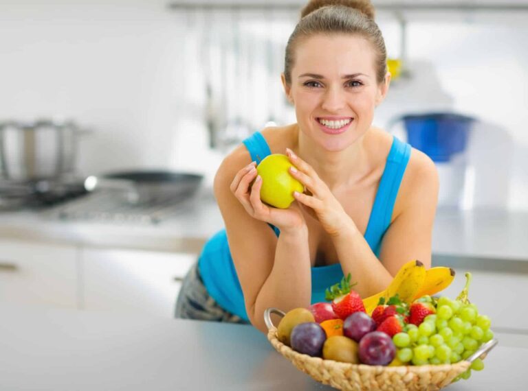 A smiling woman holds a green apple with a basket of fresh fruits in front, highlighting foods good for skin health.