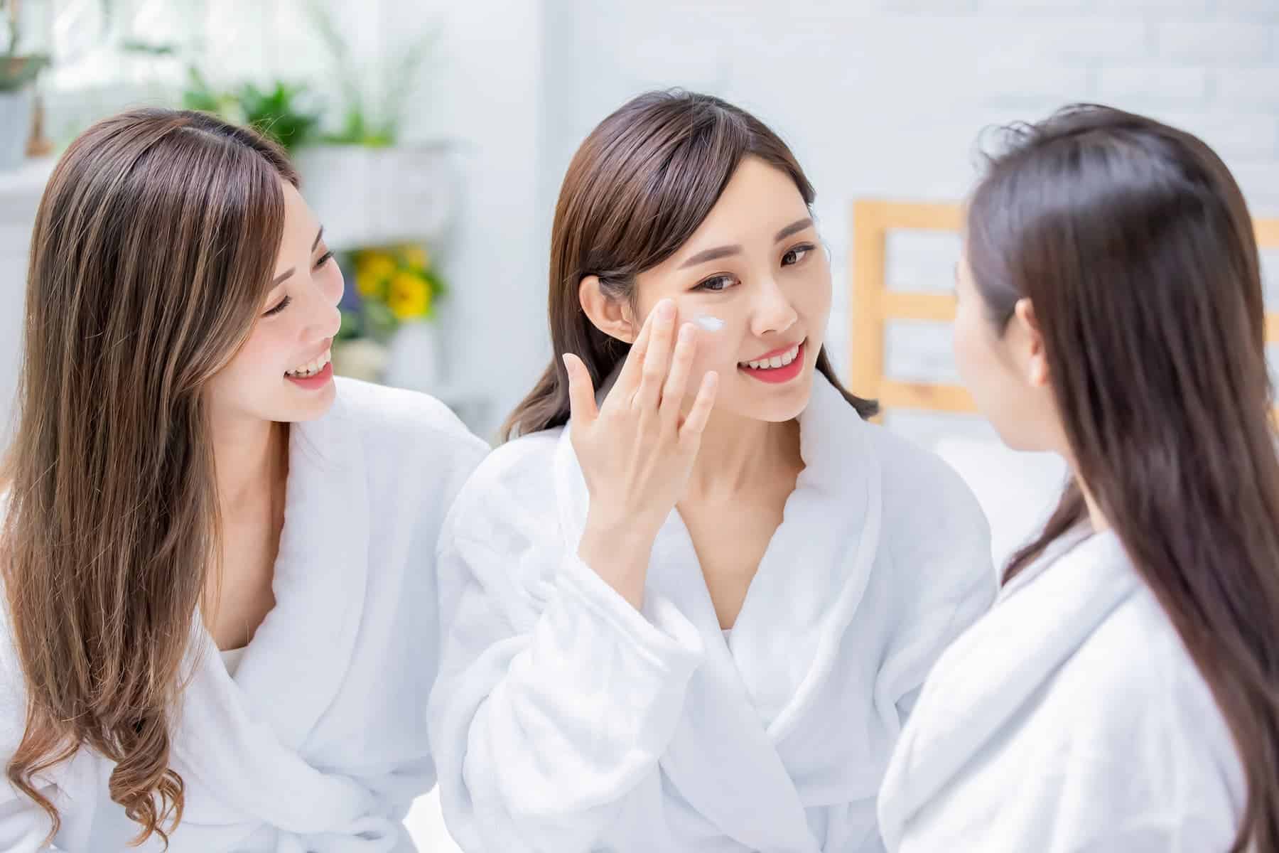 Three Korean women in white robes, with one applying cream to her face, showcasing healthy, glowing Korean skin.
