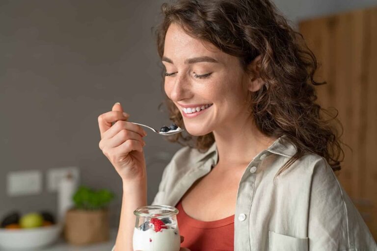 Woman smiling while eating yogurt with blueberries, enjoying a nutritious snack as part of foods for skin health.