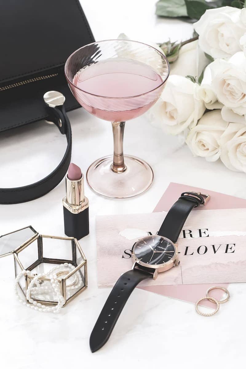A flat lay of feminine accessories including a pink cocktail, lipstick, a watch, jewelry, and roses.