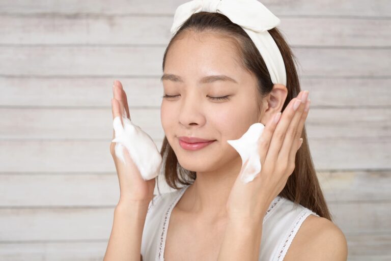 A woman with a white headband cleans her face with white foam, with her eyes closed and a gentle smile, highlighting best Korean skincare.