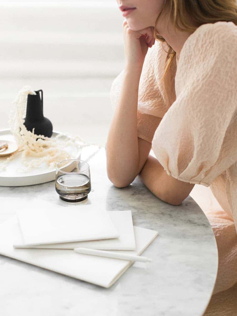 A woman in a light pink dress sits at a marble table with a notebook and glass of water, reflecting a calm and intentional lifestyle.