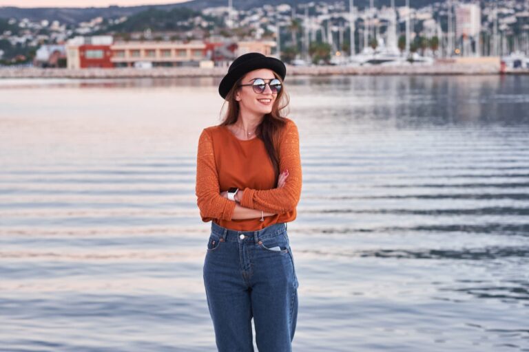 A woman in a black hat and sunglasses stands near a harbor with her arms crossed, wearing an orange top and blue jeans.