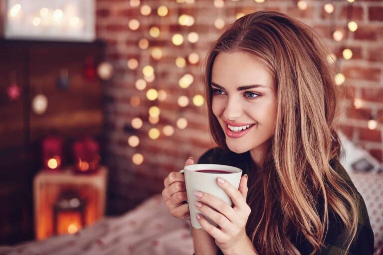 A smiling woman holds a mug of a dark beverage with blurred fairy lights and a brick wall in the background.