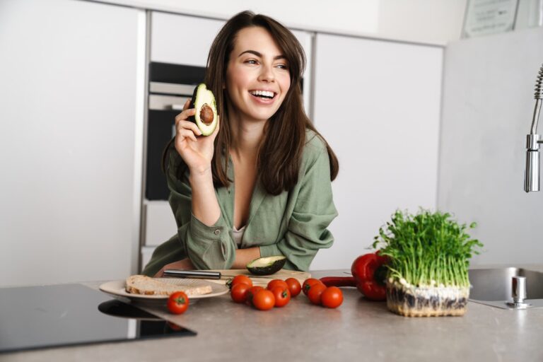 A woman laughs while holding a piece of avocado in a kitchen with vegetables on the counter.