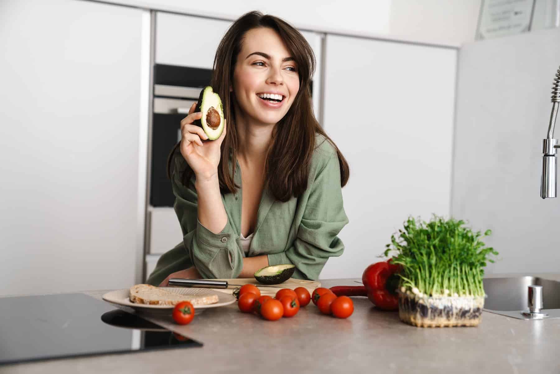 A woman laughs while holding a piece of avocado in a kitchen with vegetables on the counter.