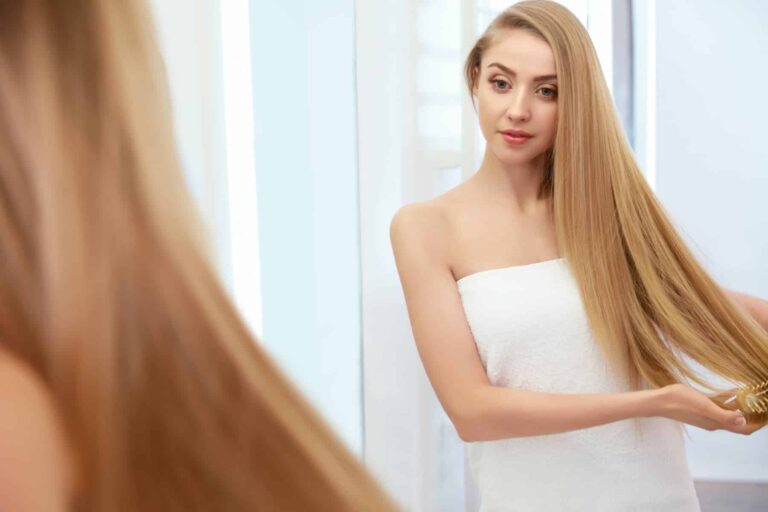 A woman with long blonde hair brushes it in front of a mirror while wrapped in a white towel.