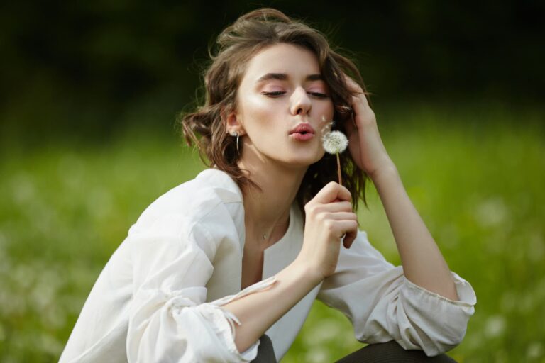 A woman wearing a white shirt sits on a grassy field and blows on a dandelion.