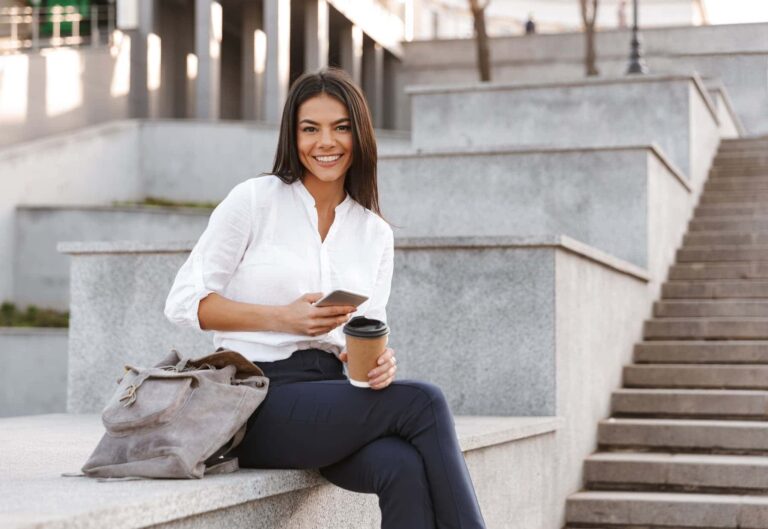 A confident woman wearing dark pants and a white shirt relaxes on stone steps while holding a phone and coffee cup.
