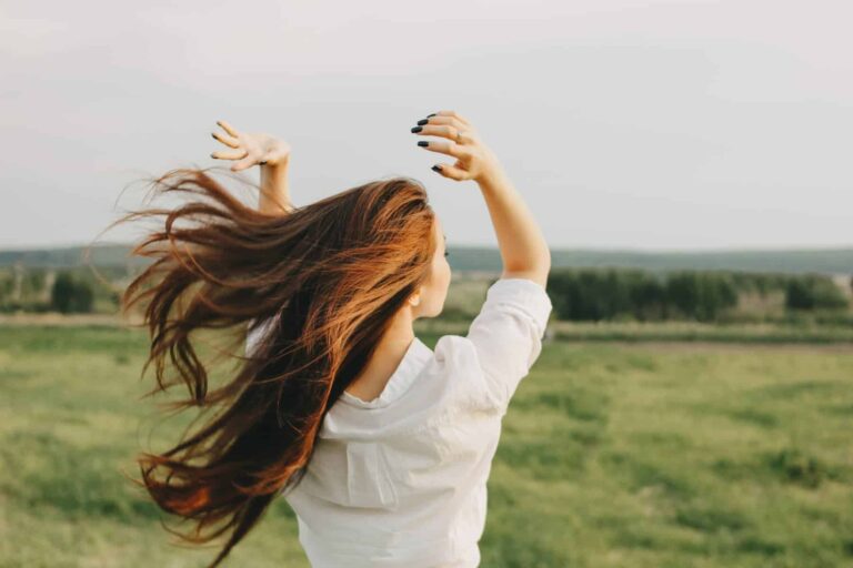 A woman with long hair stands in a field with her arms raised and her hair blowing in the wind.