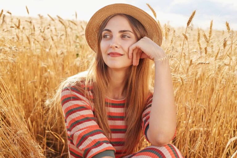 A woman in a straw hat sits among golden wheat, looking to the side while resting her chin on her hand.