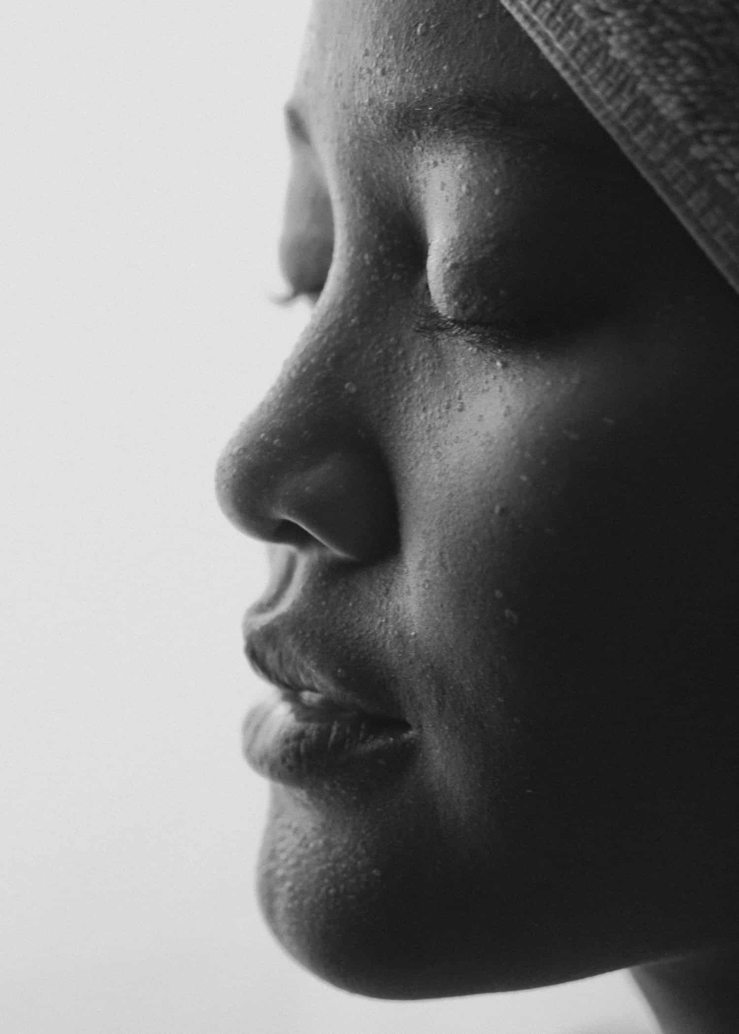 A close-up, black and white profile of a woman's face, featuring visible water droplets on her skin.
