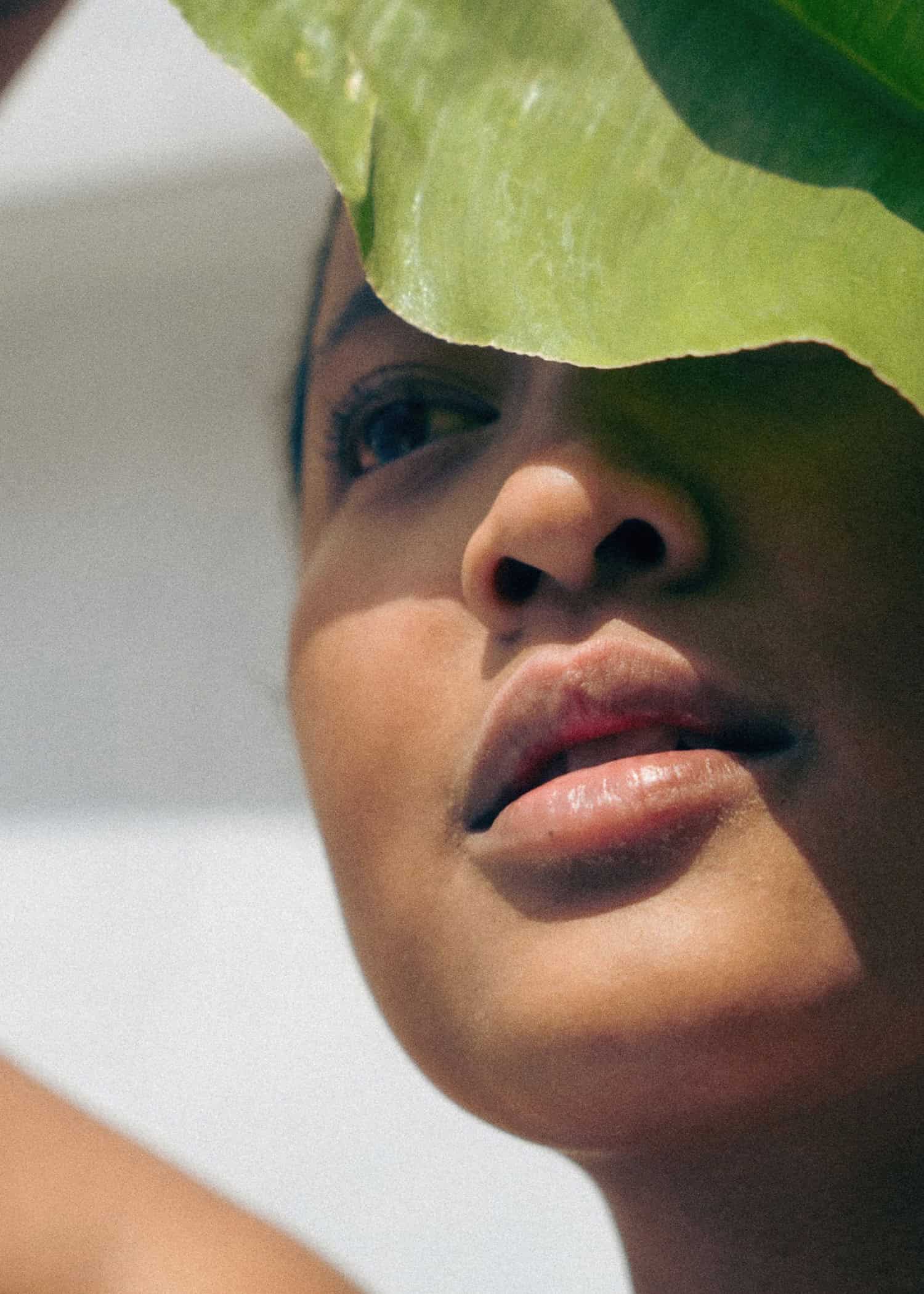 A close-up portrait of a woman's face, partially obscured by a vibrant green leaf.