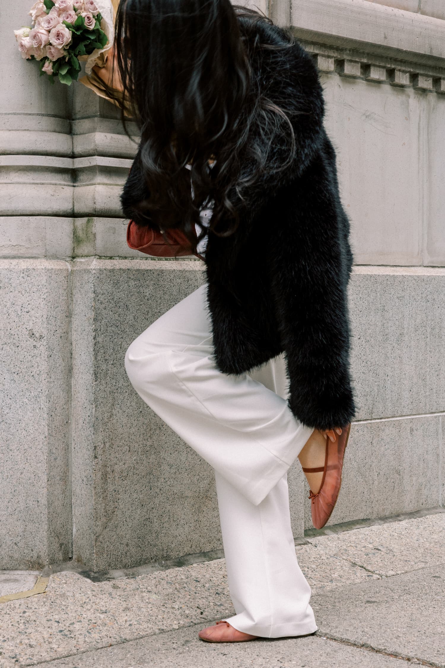 Woman in fur coat and white pants holding flowers pauses to fix her shoe while walking in the city.