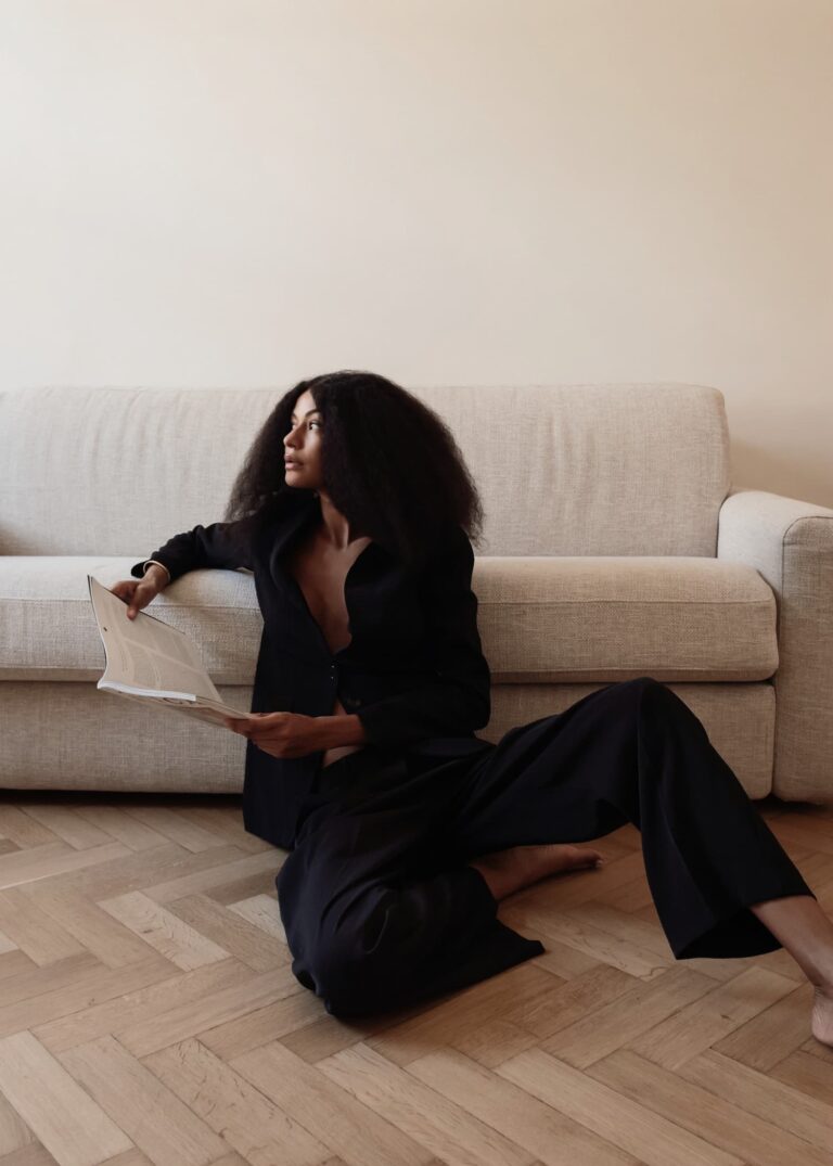 A stylish woman with voluminous dark hair sits barefoot on a herringbone floor, holding a magazine.