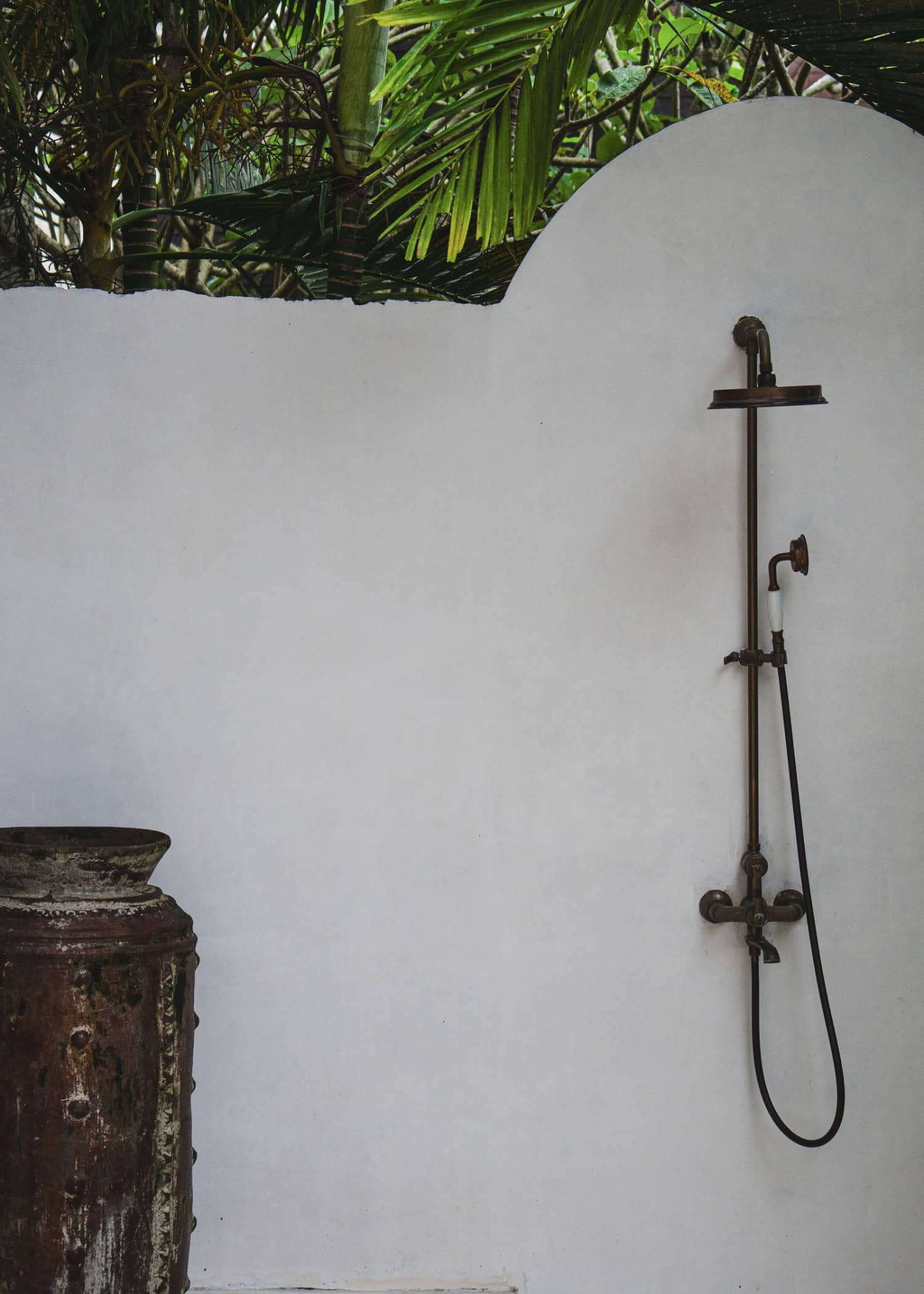 An outdoor shower with vintage brass fixtures is mounted on a textured white wall.